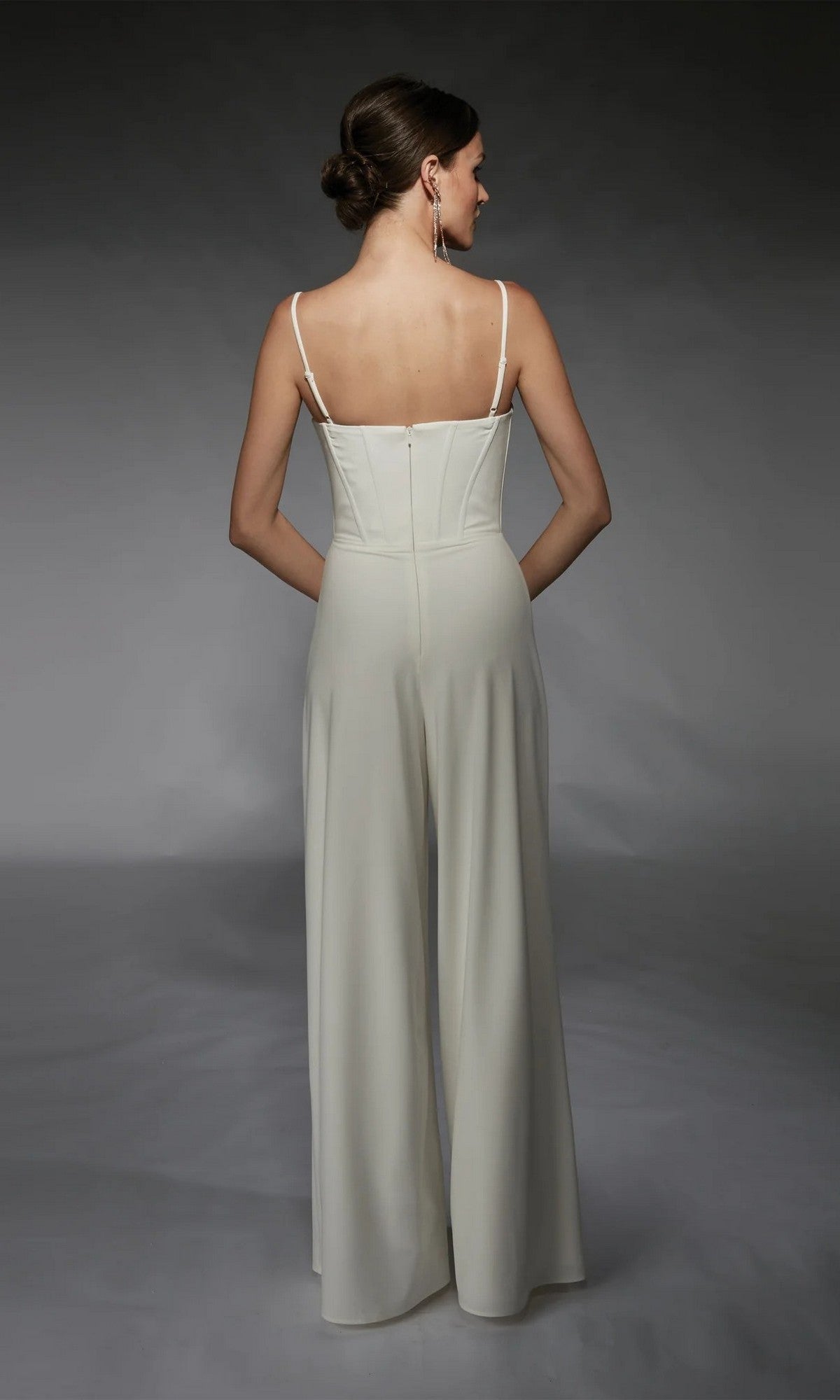 A woman wears the Alyce 70075 Long Formal Jumpsuit, featuring a sleeveless, corset-boned bodice and wide legs. She styles her hair in a low bun and completes the look with long earrings.